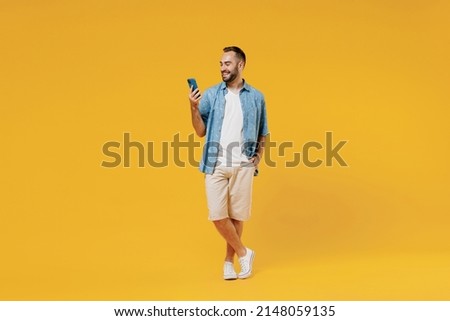 Full body young smiling happy cool caucasian man 20s wearing blue shirt white t-shirt hold in hand use mobile cell phone isolated on plain yellow background studio portrait. People lifestyle concept