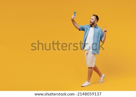Full body young happy man 20s wearing blue shirt white t-shirt doing selfie shot on mobile cell phone post photo on social network show v-sign walk isolated on plain yellow background studio portrait.