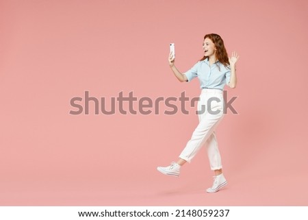 Full length side view of young friendly redhead woman in blue shirt pants doing selfie shot on mobile phone talk by video call waving hand greeting isolated on pastel pink background studio portrait