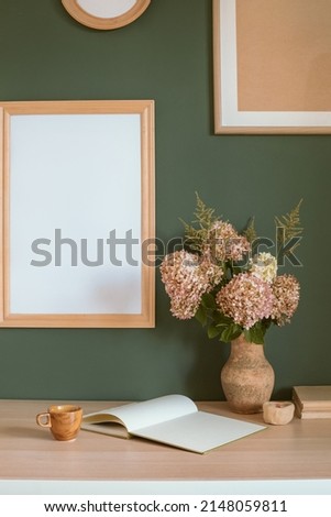 Various wooden picture frames mock ups on the wall, ceramic vase with hydrangea flowers, books on wooden table. Home office desk.