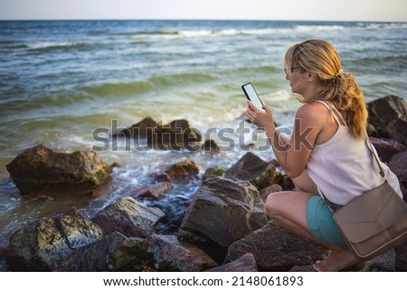 Beautiful happy young woman in mild light clothes with a careful braid, takes pictures of the landscape of the sea horizon on a modern phone while sitting on wet stones