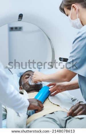 Nurse putting oxygen mask on face of patient lying with closed eyes on MRI table in procedure room