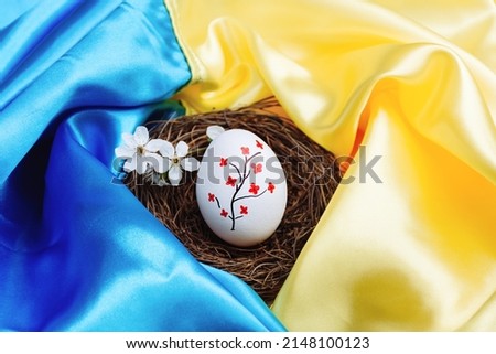 White Easter egg with beautiful painted red flowers in a wooden wicker nest on a blue and yellow bright satin background of the State Flag of Ukraine. Support for the country during the occupation.