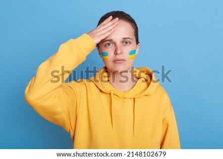 Portrait of sad woman in yellow hoodie with Ukrainian flag on cheeks, keeping her hand on head, worries about his country, posing isolated over blue background.
