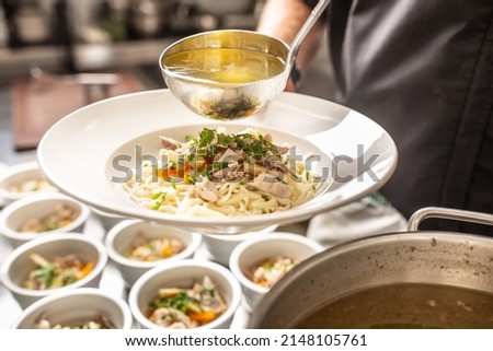 Chef pours broth into a chicken soup with noodles, meat and vegetable.
