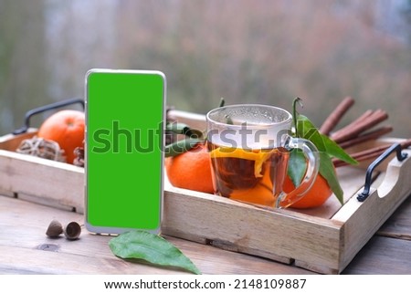 mobile phone with green screen, glass of hot tea on tray, delicious vitamin tangerines with leaves, cinnamon sticks, pine cones, cozy mood, concept vacation, weekend in nature, breakfast on terrace