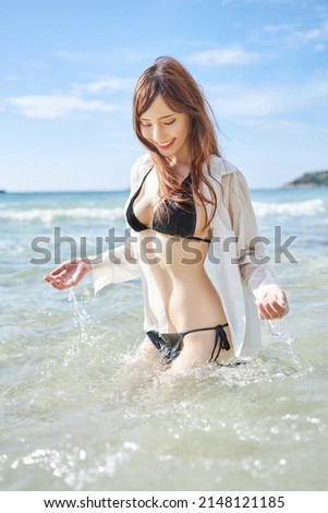 Beautiful smiling mixed race Asian and Caucasian young lady with white skin in black bikini and white shirt splashing and playing in summer sea water 