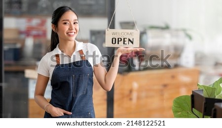 Young female barista owner coffee shop standing at her shop with open sign board, smiling and looking at camera, small business.