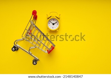 Alarm clock on the shopping cart on yellow background. Shopping time concept.