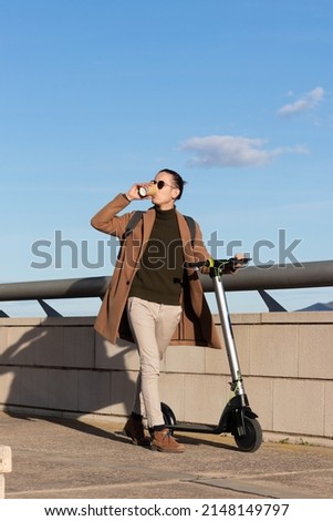 Young bussinesman drinking coffee on a cup while scrolling around the city with his electric scooter under a blue sky
