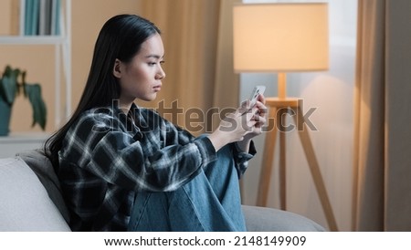 Side view of sad bored Asian woman girl sitting on sofa at home with telephone serious unhappy lady scrolling browsing in smartphone and using mobile phone app watching social media news feed typing Royalty-Free Stock Photo #2148149909