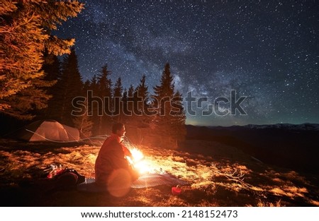 Side view of alone hiker, warming himself by campfire on mat, looking at surrounding mountain landscapes. Luxurious starry sky, Milky Way galaxy over mountain hills.