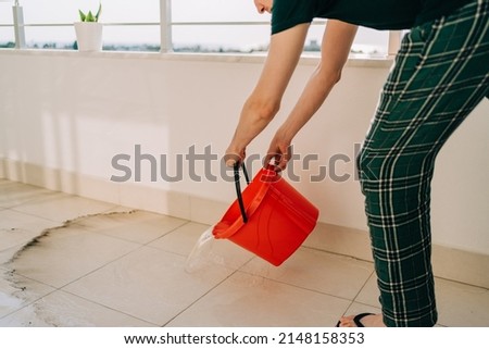 Man spilling the water from red bucket onto the tiled floor on the balcony in sunny day. Adult male doing home chores washing floors on patio. Plant in a flower pot standing on balcony railings. Royalty-Free Stock Photo #2148158353