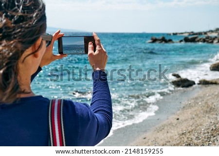 Back view of tourist woman taking photo with her smartphone of the seascape on the beach during windy spring day. Defocused foreground.