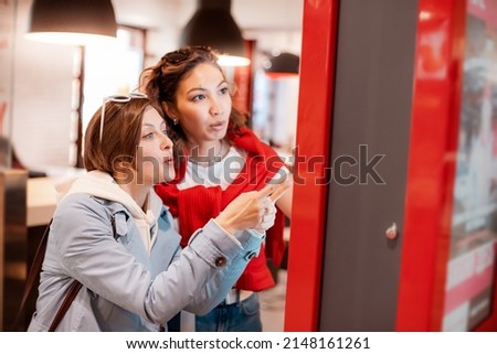 Two girl friends orders food and lunch at a fast food restaurant using a self-service kiosk or a terminal with a screen. Modern commerce equipment Royalty-Free Stock Photo #2148161261