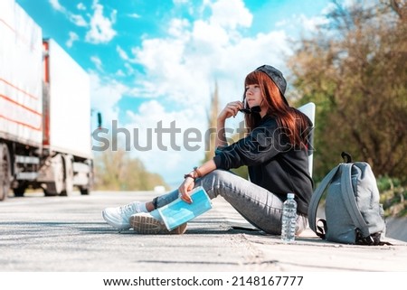 A young woman in cap with tattoed hands sitting along the road with a paper map. Copy space. Concept of hitchhiking and local travel.