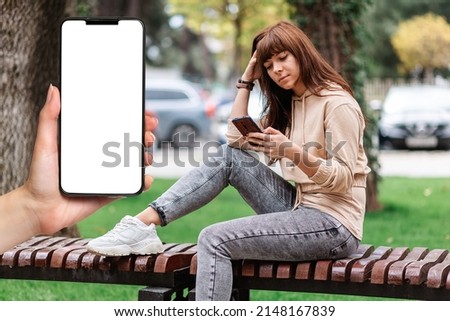 Free wi-fi zone. A woman's hand holds a smartphone with a white screen, close-up. A young woman sits on a bench and uses a smartphone. The concept of online applications for the smartphone.