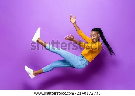 Full body portrait of joyful African American woman in causal wear falling and levitating on purple background in light studio