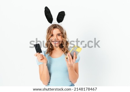 Joyful young woman wearing Easter Bunny ears costume holding smartphone and plastic bank card standing on white background