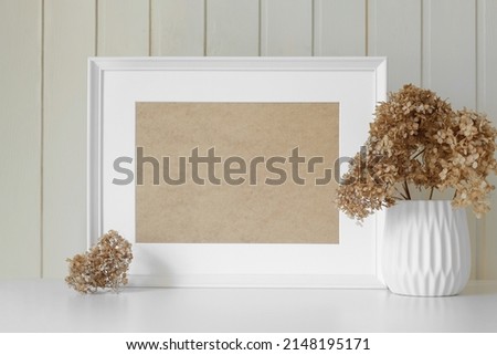Empty picture frame with beautiful bouquets of dried hydrangea flowers. Modern white ceramic vase with dry grass. Wooden wall background. Elegant Scandinavian interior. Summer, fall still life photo.
