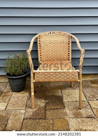 Single tan woven rattan chair sitting on a concrete block patio with a potted rosemary bush in front of a dark pacific blue vinyl plank house.
