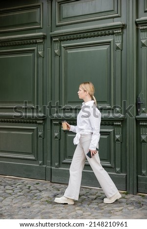 full length view of trendy woman with smartphone and coffee to go walking along grey wall