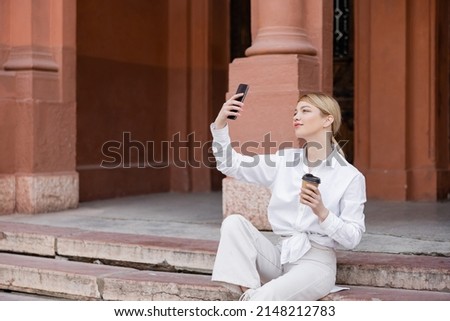 pretty woman with coffee to go sitting on stairs and taking selfie on cellphone