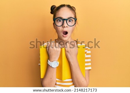 Beautiful brunette little girl wearing glasses and holding book afraid and shocked with surprise and amazed expression, fear and excited face.  Royalty-Free Stock Photo #2148217173