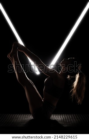 Fit woman practicing yoga poses. Silhouette girl doing exercise in studio against black background with v shaped white led tube light. No stress inner balance concept. Royalty-Free Stock Photo #2148229615