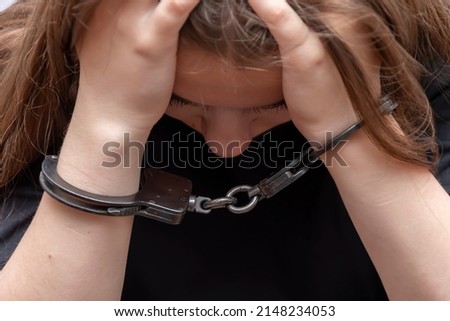 A young girl handcuffed hides her face on a gray background, close-up. Juvenile delinquent in a black T-shirt, criminal liability of minors. Royalty-Free Stock Photo #2148234053