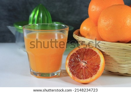 Glass with orange juice on white table with basket full of oranges and citrus squeezer next to it