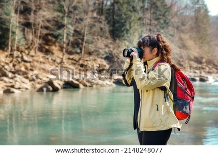 Photographer woman near a mountain river standing on the rock. Female enjoying the moment of the nature in sunny day. Woman is holding photo camera and taking photo of the view.