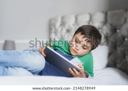 Young boy using tablet playing game on internet with friends, Homeschooling Kid doing homework online by digital pad at home, Child lying on bed relaxing,watching cartoon or talking online with friend