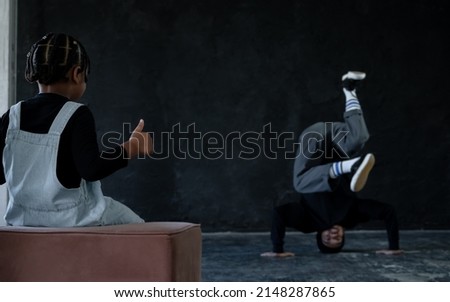 Little African kid boy thumbs up while looking at Asian young man with tattoo dancing hip hop dance stand on head on black background. Cool man teacher showing his passionate in dancing to student