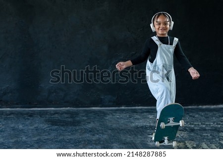 Portrait of smiling African American little kid boy with skateboard on black background. Happy adorable child wear white headphones, bib jeans, necklace chain in hip hop style