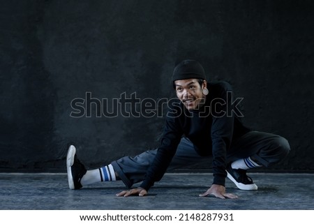 Asian hip hop young man with tattoo stretching exercise his legs on floor before dance on black background. Attractive man with moustache smiling wear knitted hat, ear piercing warm up body