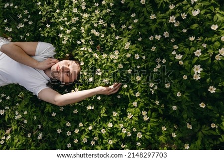 The portrait of beautiful cute woman no makeup in white shirt laying down on green grass with white flowers. Sunny summer day, wild nature, mood vacation, relax. Top view of girl. Be free, love forest