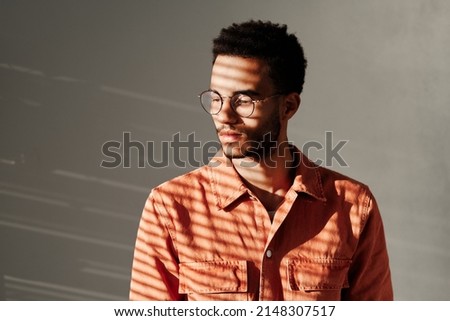 Calm young mixed race man in round-shaped glasses standing under light through blinds, isolated background