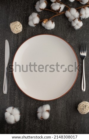 Empty white dish with knife and fork on a slate background with a cotton branch, with copy space for your menu or recipe. Menu card for restaurants and table setting. Vertical photo. Flat lay