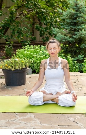 Young woman practices yoga in the garden, sitting in the Lotus position, hands folded in Jnana mudra and meditating.