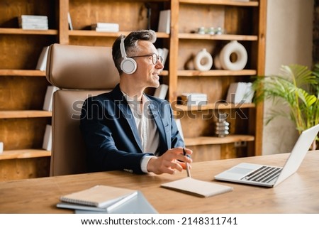 Caucasian mature middle-aged businessman ceo freelancer boss employee having break after hard-working day in office, relaxing while listening to music radio podcast in headphones at the desk