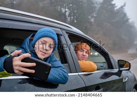 Happy father and son Sitting together in a new car on a journey. Family are resting on the side of the road on a road trip. Child takes pictures on smartphone. Happy family travels.