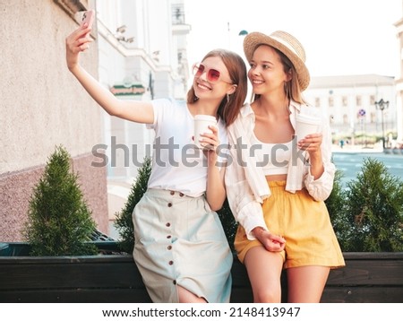 Two young beautiful smiling hipster female in trendy summer clothes.Sexy carefree women posing in the street. Positive pure models taking selfie. They drinking coffee or tea in plastic cup Royalty-Free Stock Photo #2148413947