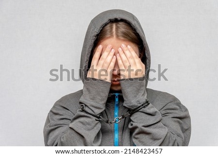A teenage girl is handcuffed, hiding her face, in a hoodie, on a gray background. Juvenile delinquent, criminal liability of minors. Members of youth criminal groups and gangs. Royalty-Free Stock Photo #2148423457