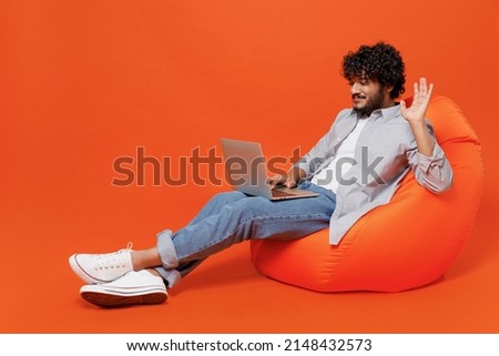 Full size young bearded Indian man 20s years old wears blue shirt sit in bag chair get video call using laptop pc computer talking greet with hand isolated on plain orange background studio portrait