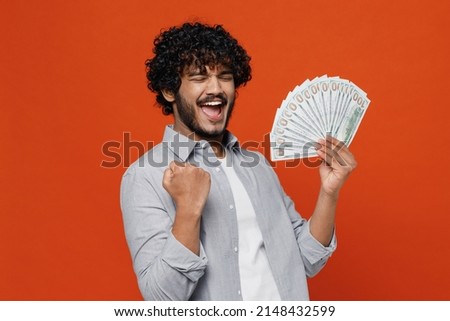 Young bearded Indian man 20s years old wears blue shirt holding fan of cash money in dollar banknotes doing winner gesture celebrate clenching fists isolated on plain orange background studio portrait Royalty-Free Stock Photo #2148432599