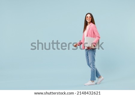 Full length side view young smiling happy woman 20s in pastel pink clothes glasses hold closed laptop pc computer walk isolated on blue background studio portrait. People lifestyle technology concept