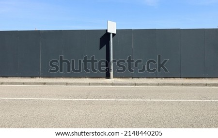 Black wooden panels fence and a pole with a blank sign. Concrete sidewalk and asphalt road in front, sky above. Background for copy space.