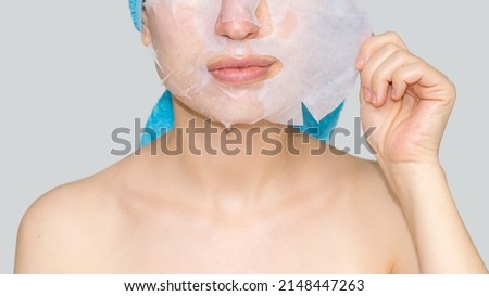woman with blue towel on head removing face mask sheet with one hand. anti wrinkle, moisturizing. mock up,grey background space for text. bottom part of the face, mouth, neck, chest and nose visible.