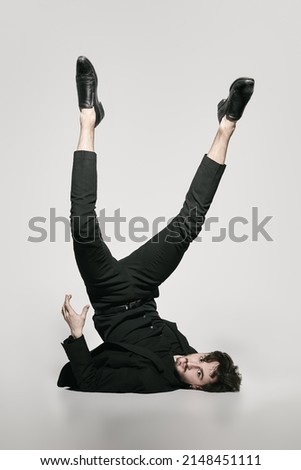 Fashion and Art. A stylish brunet man fashion model in an elegant black suit poses on a white background upside down with his legs up. Full-length studio portrait. Royalty-Free Stock Photo #2148451111